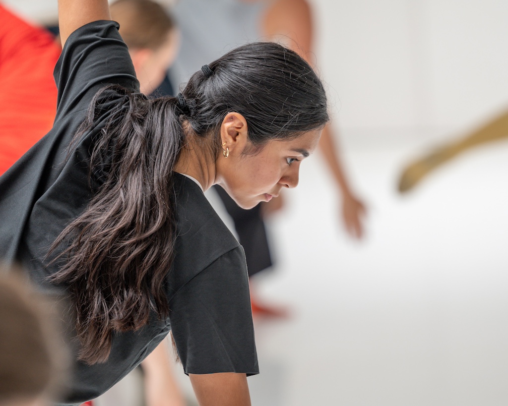 A dancer looks down while extending both arms during a training session in a dance studio